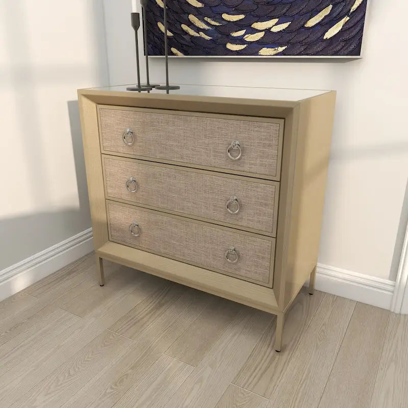 Commode en bois à trois tiroirs avec finition capitonnée, plateau miroir et poignées anneaux.