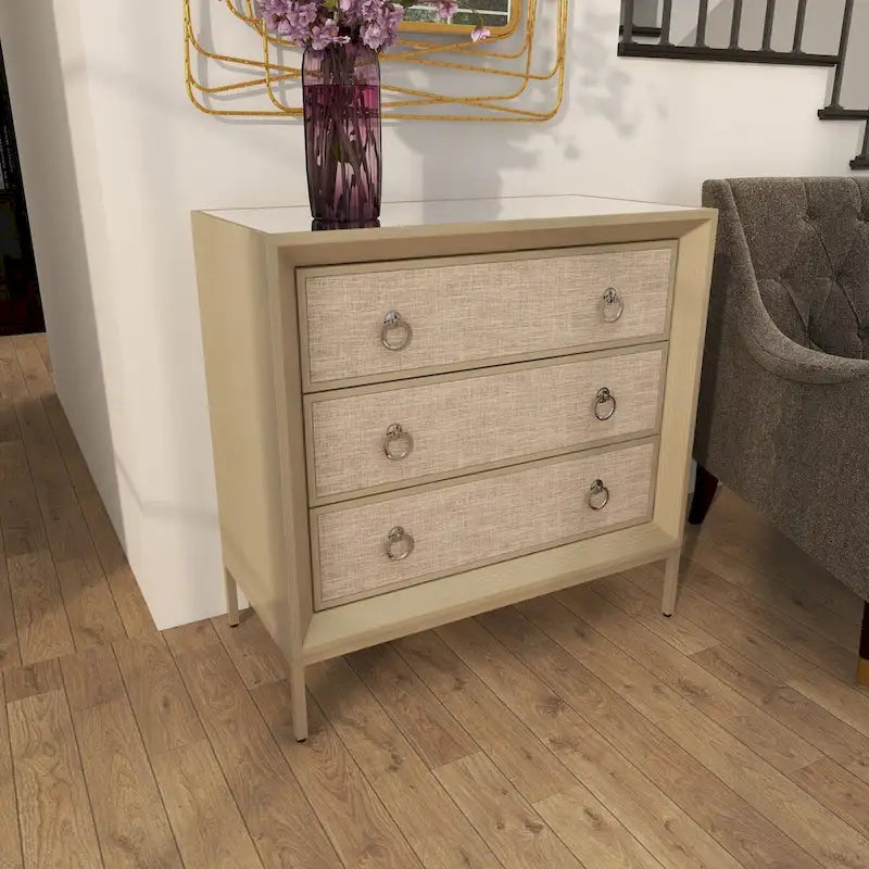 Commode en bois à trois tiroirs avec finition capitonnée, plateau miroir et poignées anneaux.