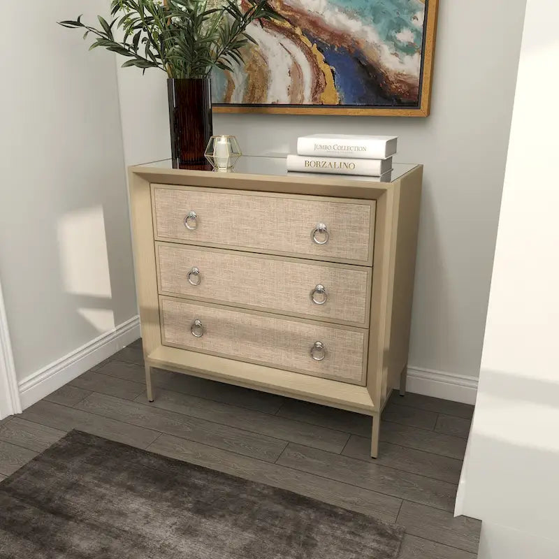 Commode en bois à trois tiroirs avec finition capitonnée, plateau miroir et poignées anneaux.